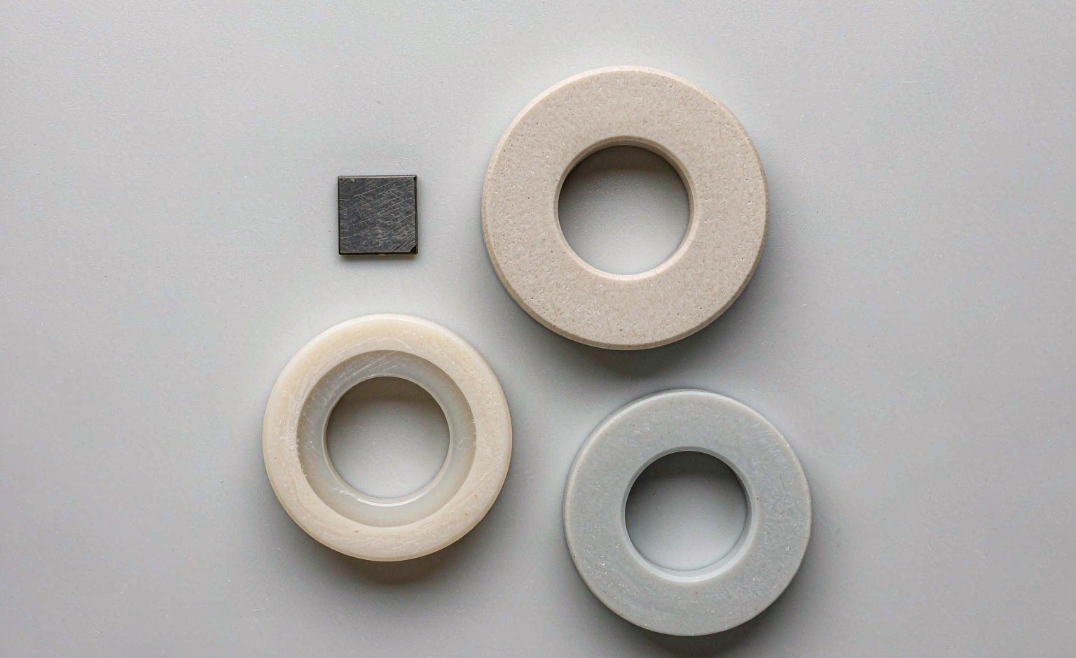 Four sintered ceramics side by side: a thin graphite-grey silicon nitride plate, an ivory alumina ring, a warm zirconia ring and a pale grey aluminium nitride disc